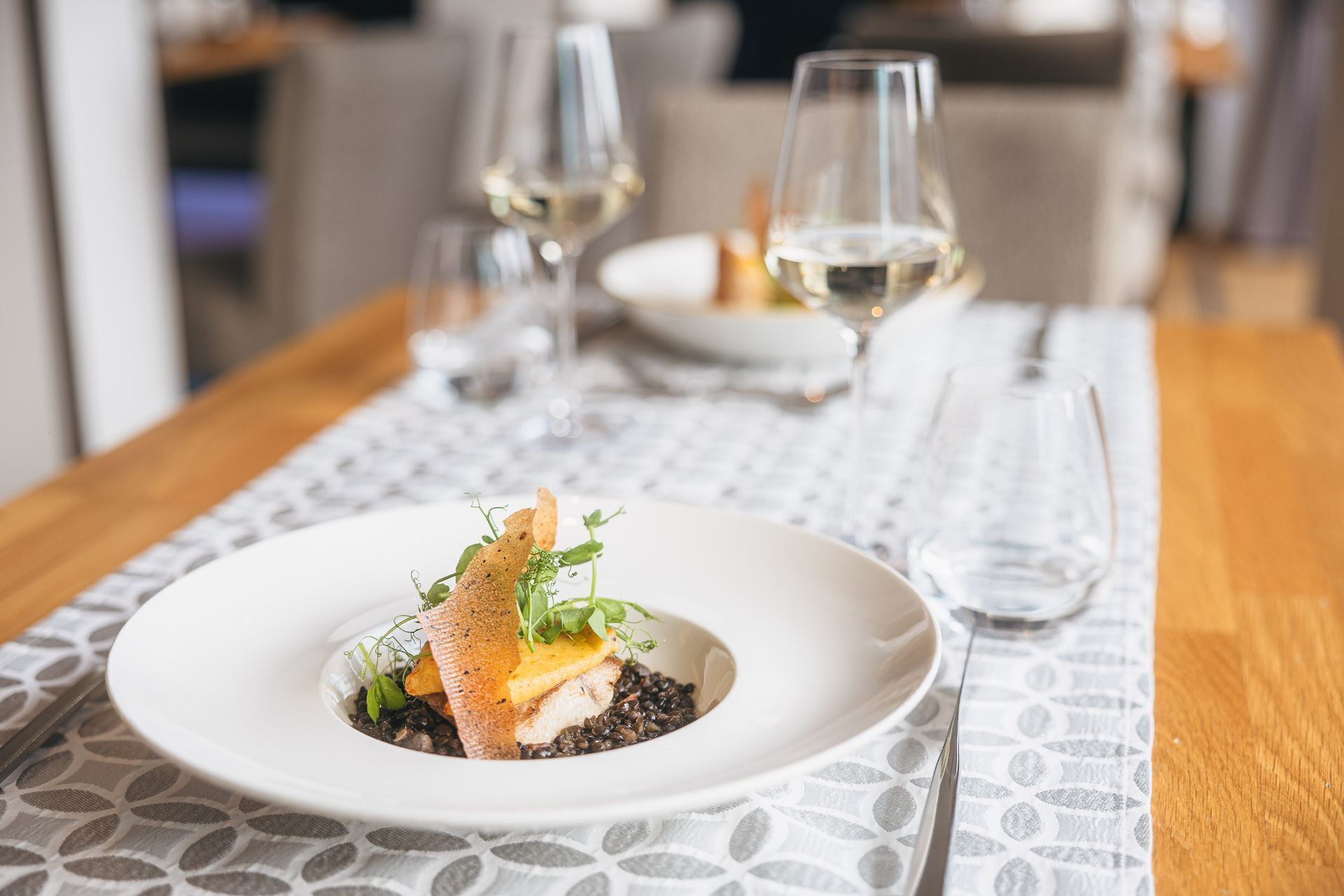 A dining table with a white plate containing a dish, a white tablecloth, and two wine glasses.