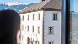 A person holding a glass of wine, looking out from a window at a building with a tower and a mountain backdrop.