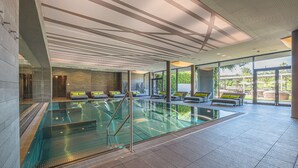 A modern indoor swimming pool with glass walls, multiple lounge areas with green cushions, and a view of the outdoors.