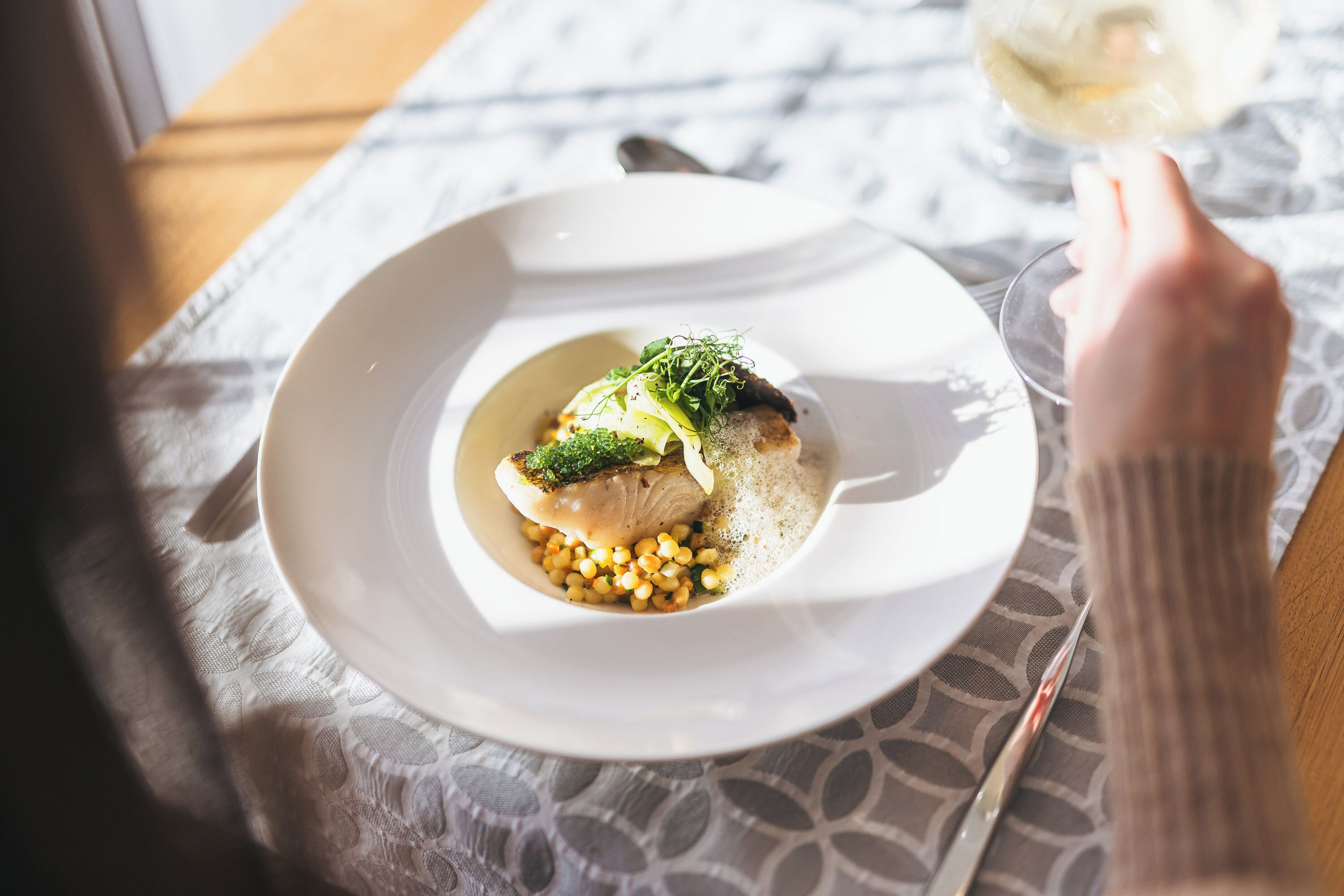 Une assiette blanche contenant un plat de poisson, agrémenté de verdure, est posée sur une table recouverte d’une nappe à motifs.
