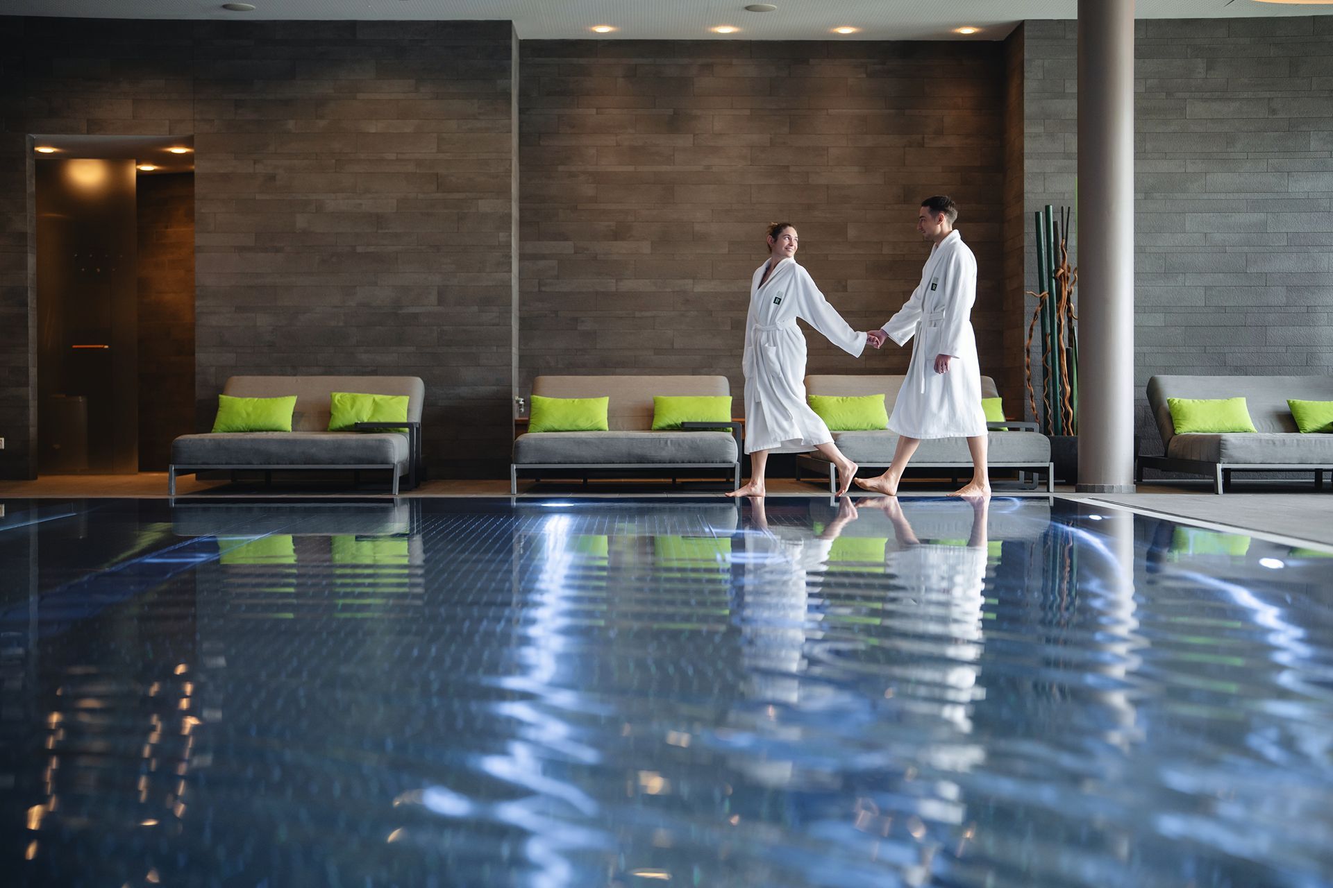 Zona de piscina con dos personas vestidas con batas blancas, sofás grises con cojines verdes y un diseño interior moderno.