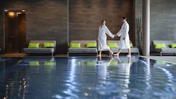 Zona de piscina con dos personas vestidas con batas blancas, sofás grises con cojines verdes y un diseño interior moderno.