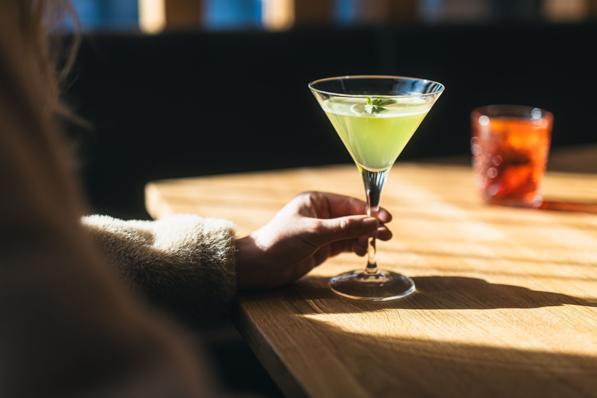 Una mano sostiene una copa de martini con una bebida verde sobre una mesa de madera.