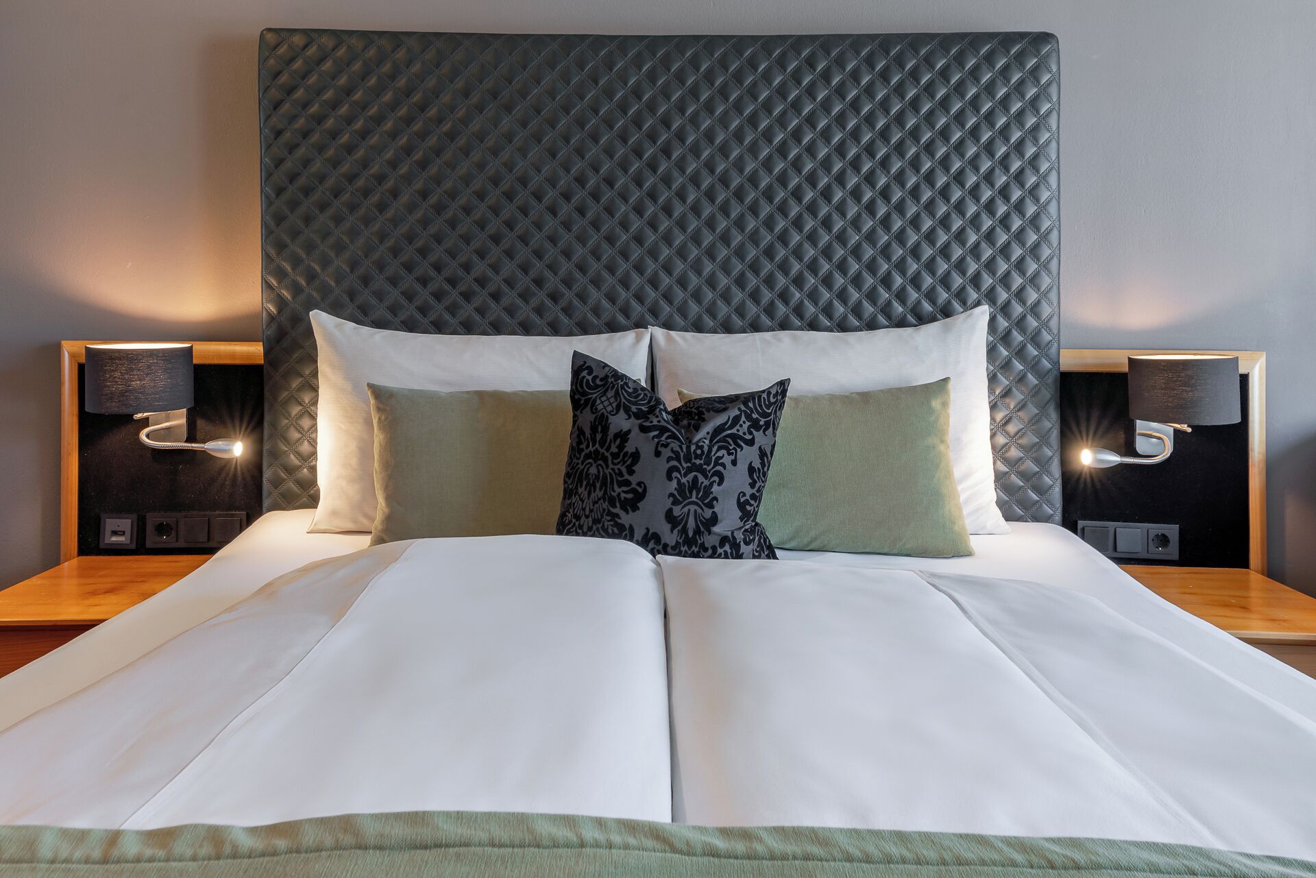 Una cama bien hecha con un cabecero acolchado, dos lámparas de noche y un plaid verde al pie de la cama.