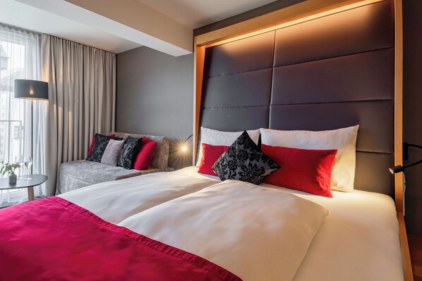 A hotel room with a large bed, a bedside table with a lamp, and a view of the outside through a window with curtains. - Ritzenhof – Hotel und Spa am See (Saalfelden am Steinernen Meer)