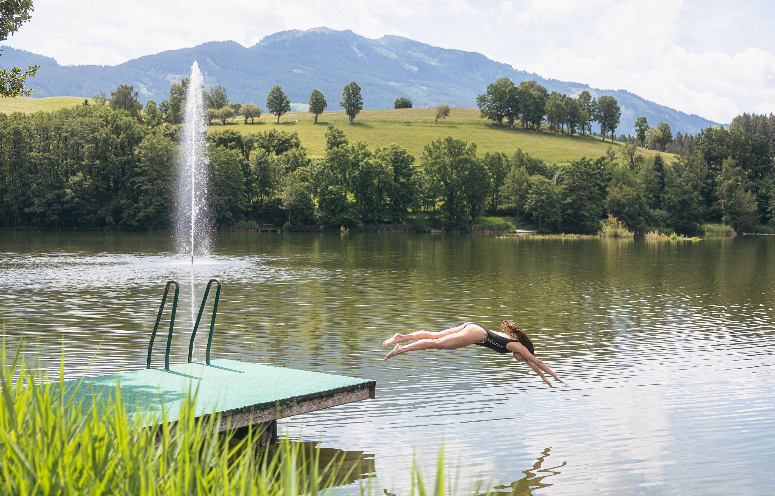 A woman diving into a lake from a dock with a fountain in the background and mountains in the distance.