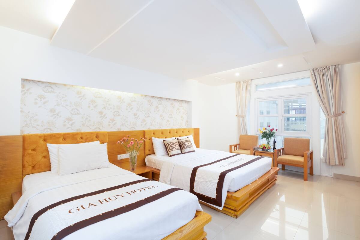 Habitación de hotel con dos camas, cabecera de madera, ventana grande con cortinas y mesita de noche con un florero.
