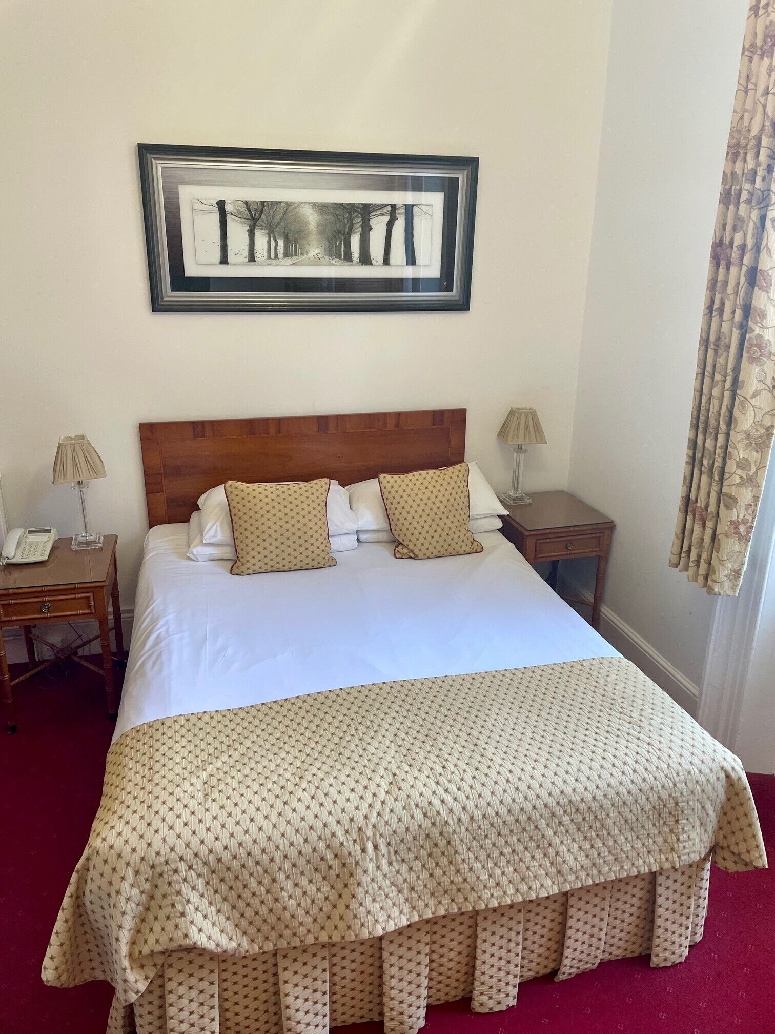 A neatly made bed with a patterned bedspread and pillows, a wooden headboard, a framed artwork above the bed, and a nightstand with a lamp on each side.