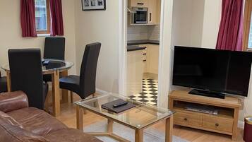 A living room with a brown leather sofa, a glass coffee table, a TV on a wooden stand, a dining area with a table and chairs, and a kitchen visible through an open door.