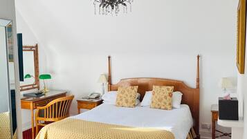 A hotel room with a large bed, a wooden desk, a chair, a desk lamp, a telephone, and a chandelier.