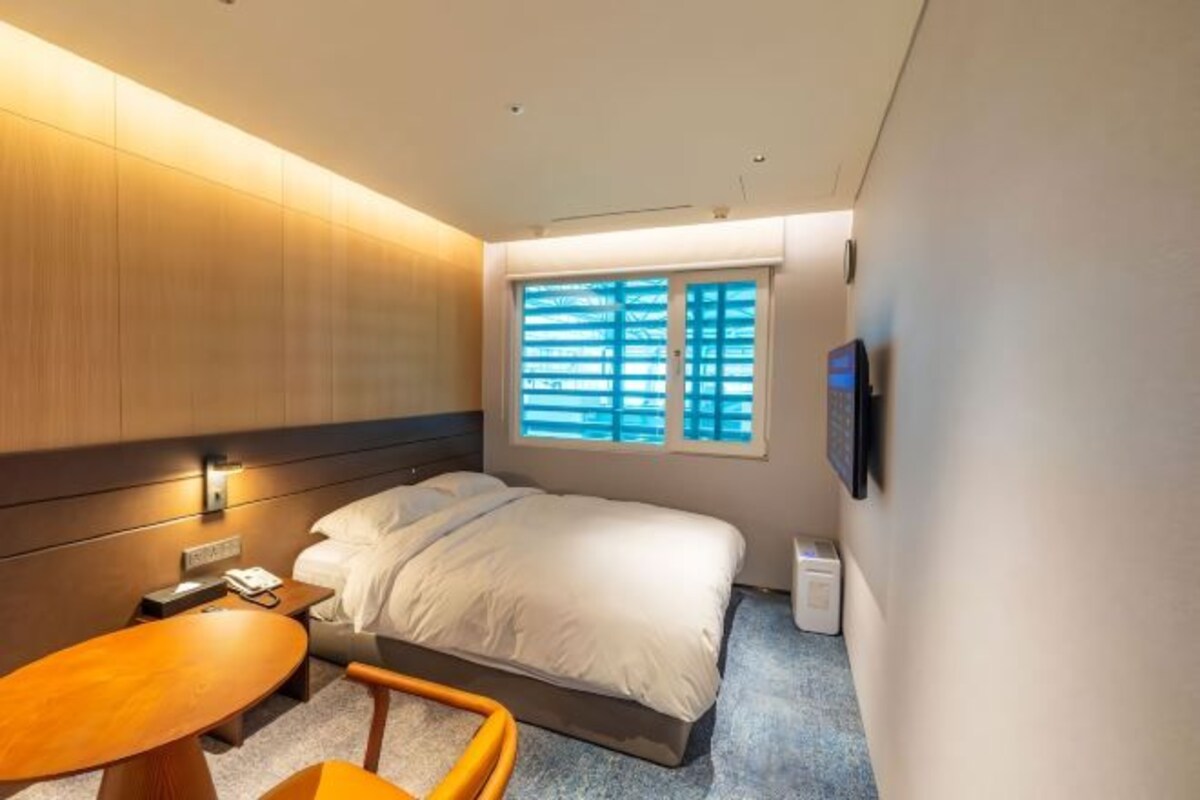 A modern hotel room with a large bed, a bedside table, a chair, a desk, and a window with blinds.
