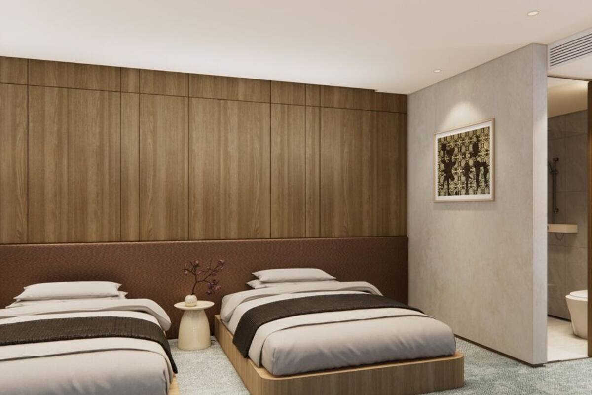 A modern hotel room with two beds, a wooden headboard, a small table with a vase, and a bathroom visible through an open door.