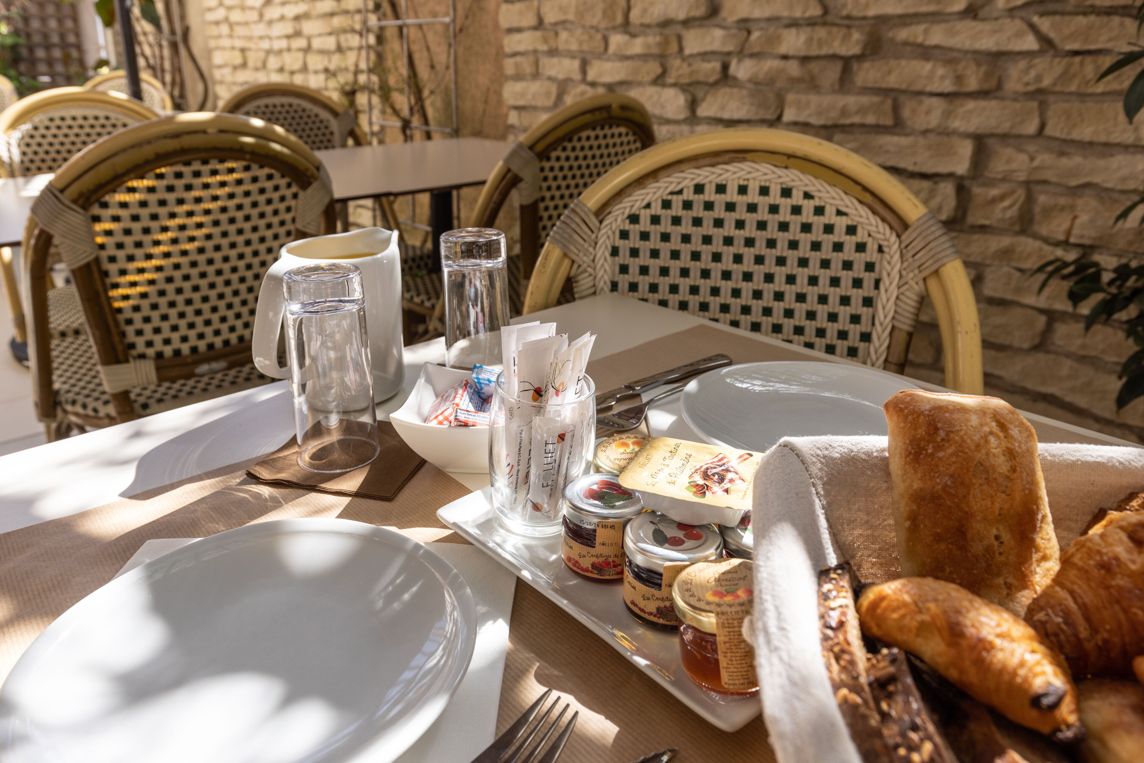 A table set for breakfast with plates, cutlery, a glass pitcher, jars of jam, and bread.