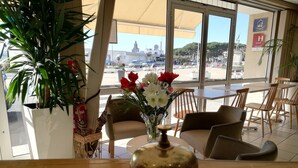 Zona d'entrada d'hotel amb un taulell de recepció de fusta, una campana i un vas amb flors. Hi ha cadires i taules disposades per als clients, i es veu el carrer a l'exterior.