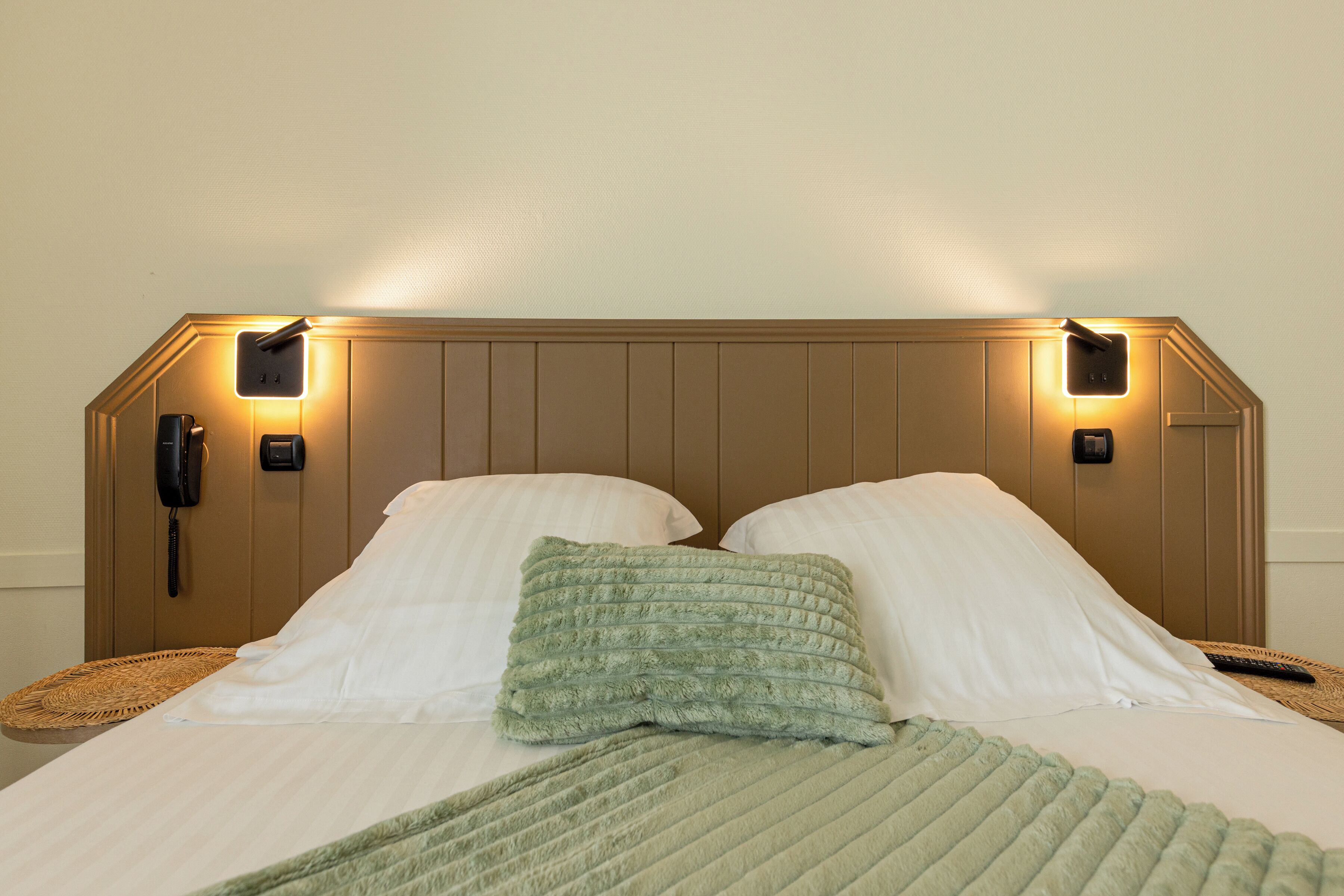 Una cama con sábanas blancas, una colcha verde y dos almohadas. Hay dos lámparas de pared a cada lado de la cama.