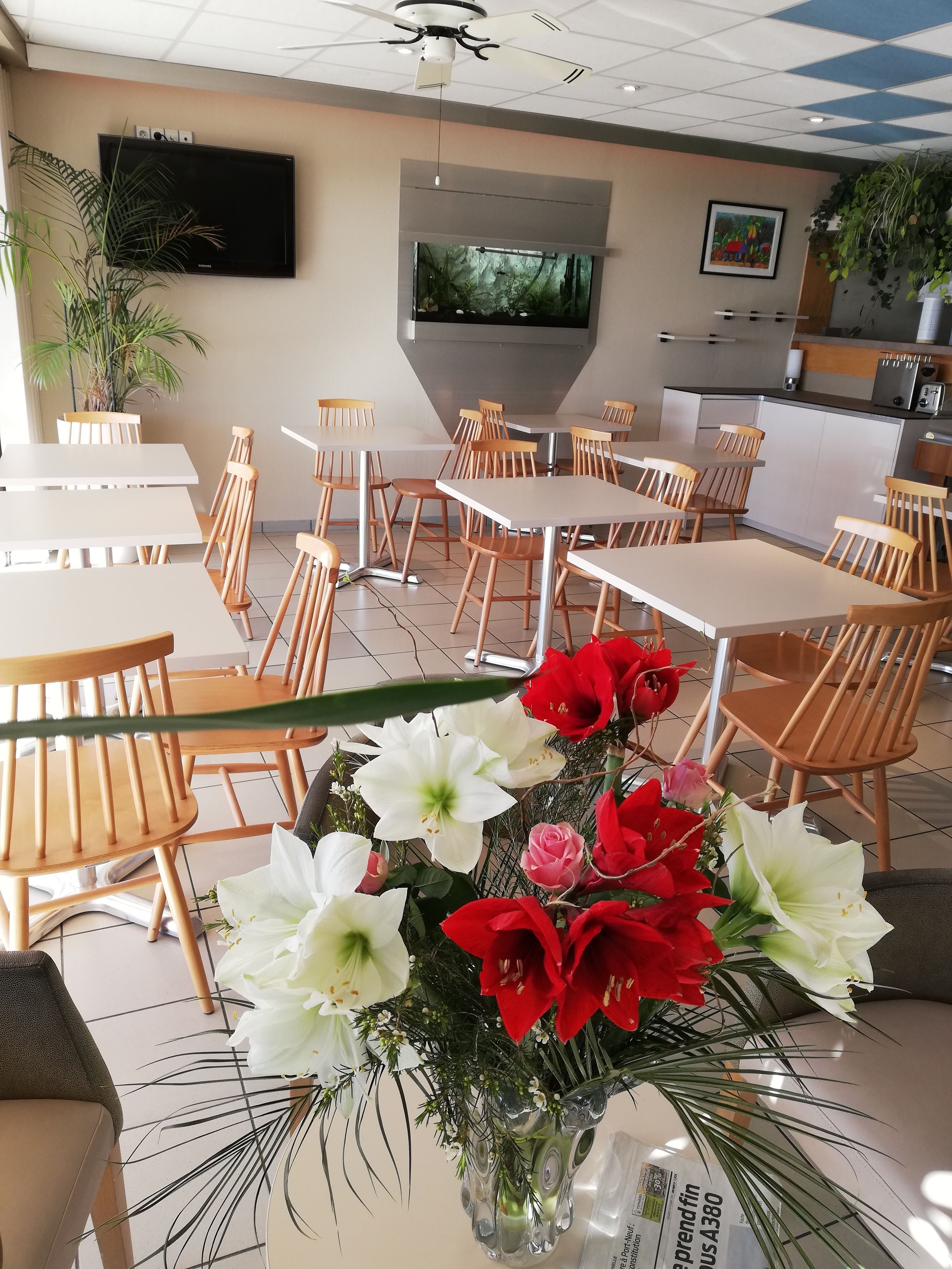 A hotel dining area with tables and chairs, a flat-screen TV on the wall, and a kitchen in the background.