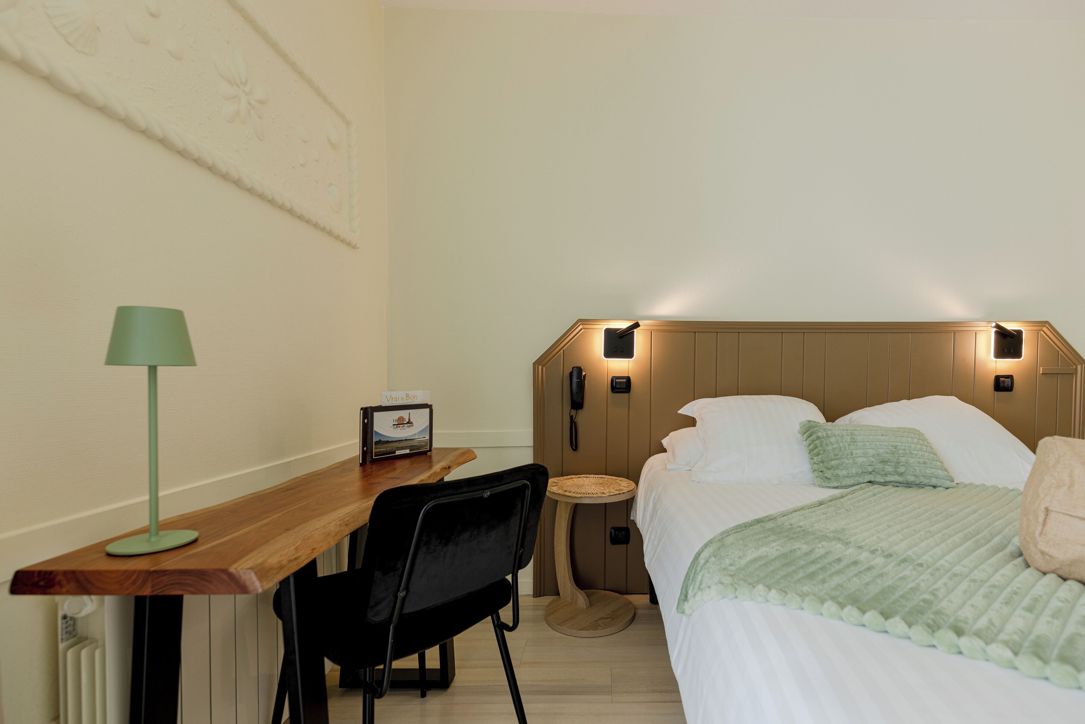 A hotel room with a wooden desk, a green lamp, a bed with white bedding, a bedside table, and a wall-mounted phone.