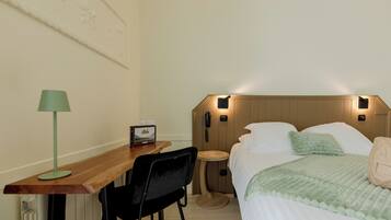 A hotel room with a wooden desk, a green lamp, a bed with white bedding, a bedside table, and a wall-mounted phone.