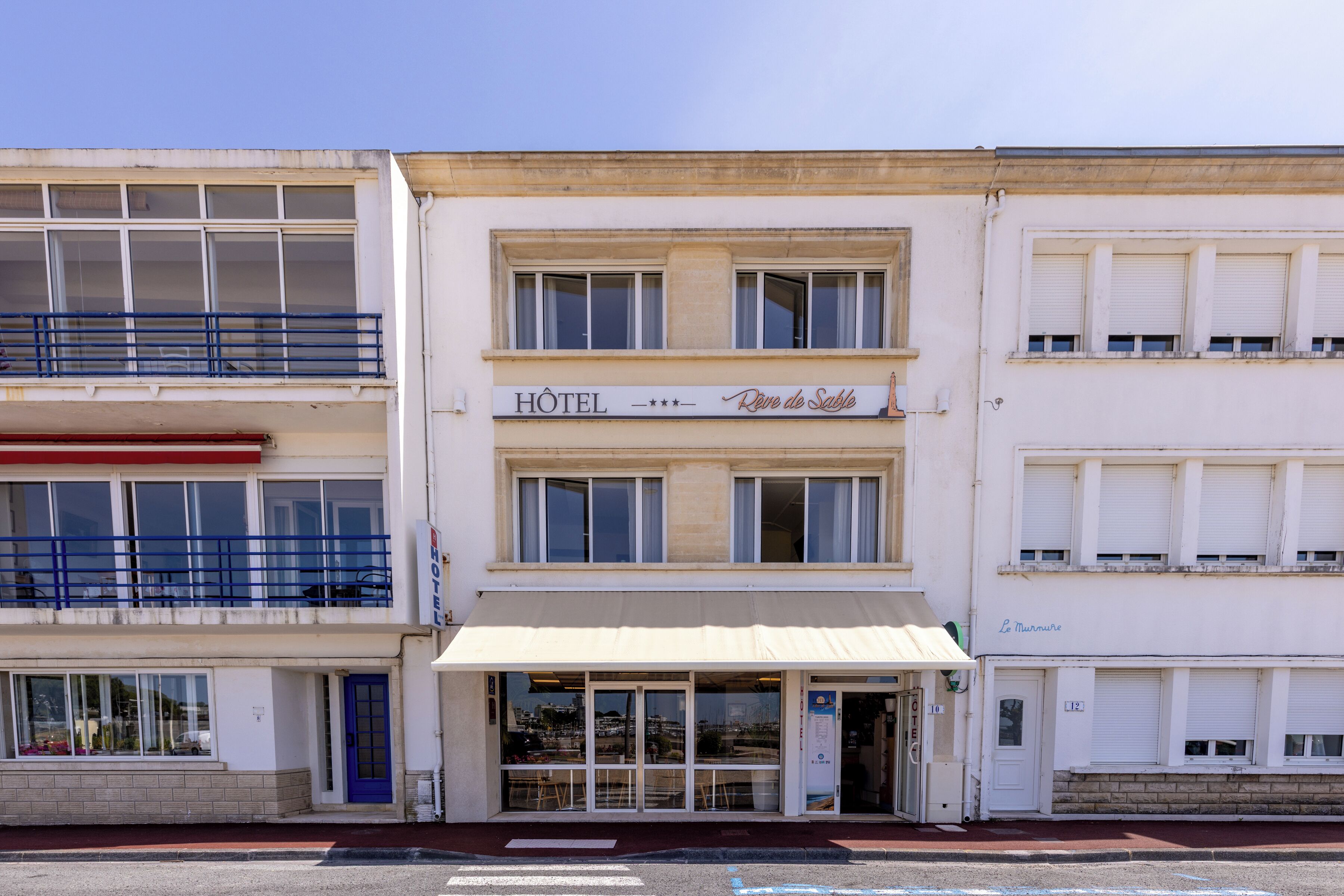 A hotel with a sign reading 'HOTEL Rive de Sable,' a balcony with blue railings, and a ground-floor entrance with a door.