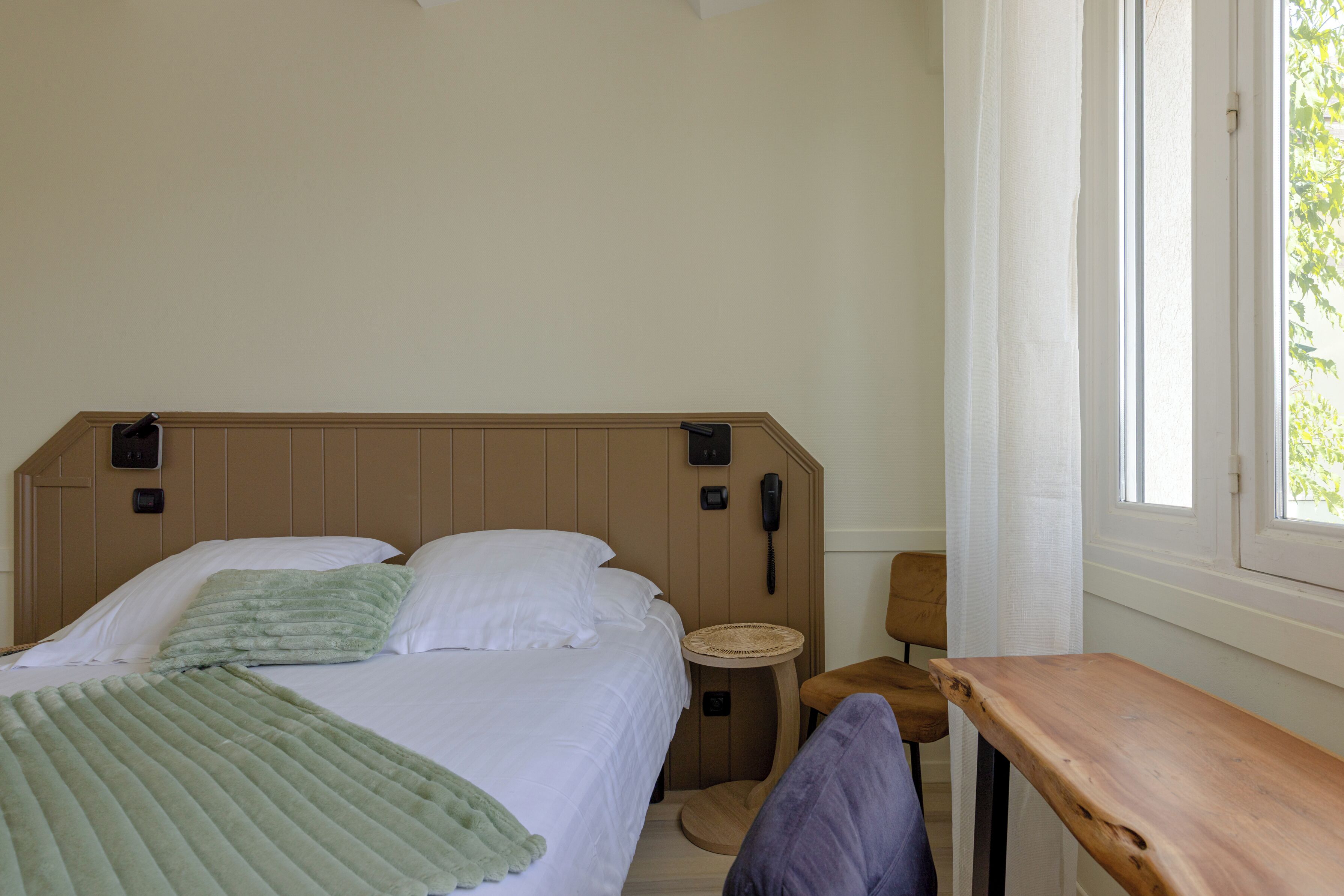Un dormitorio con cama, mesita de noche, una silla, una pequeña mesa redonda y una ventana.