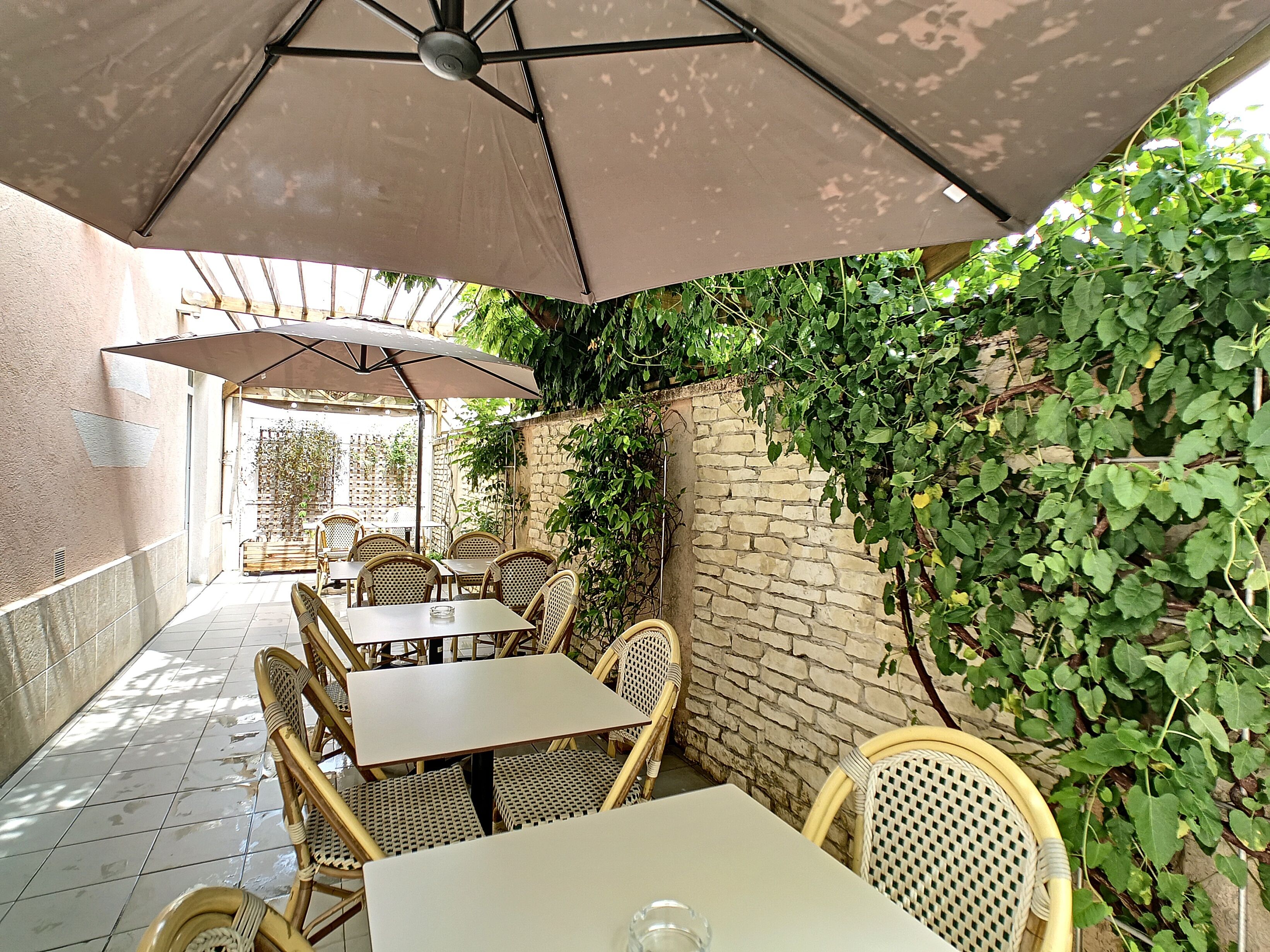 An outdoor patio with tables and chairs under umbrellas, surrounded by greenery and a stone wall.
