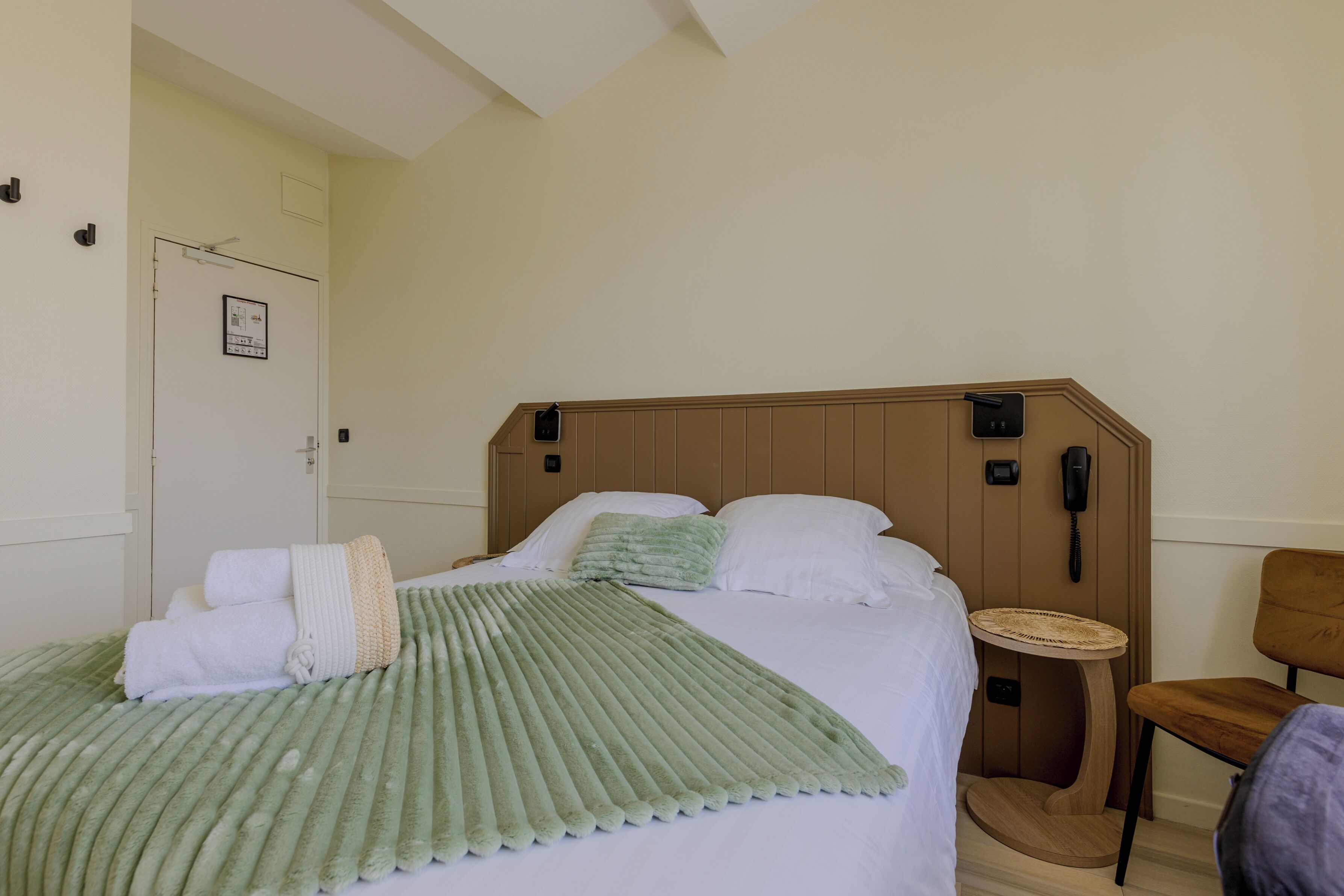 A hotel room with a bed, a bedside table, a chair, and a door with a sign.