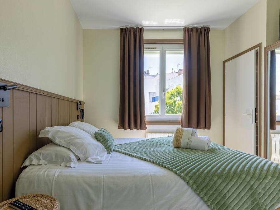 A hotel room with a bed, a telephone, a window with curtains, and a view of the outside.