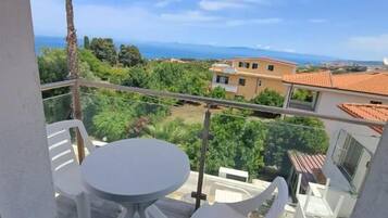 Un balcón con mesa y sillas, con vista a un paisaje costero con edificios y vegetación.