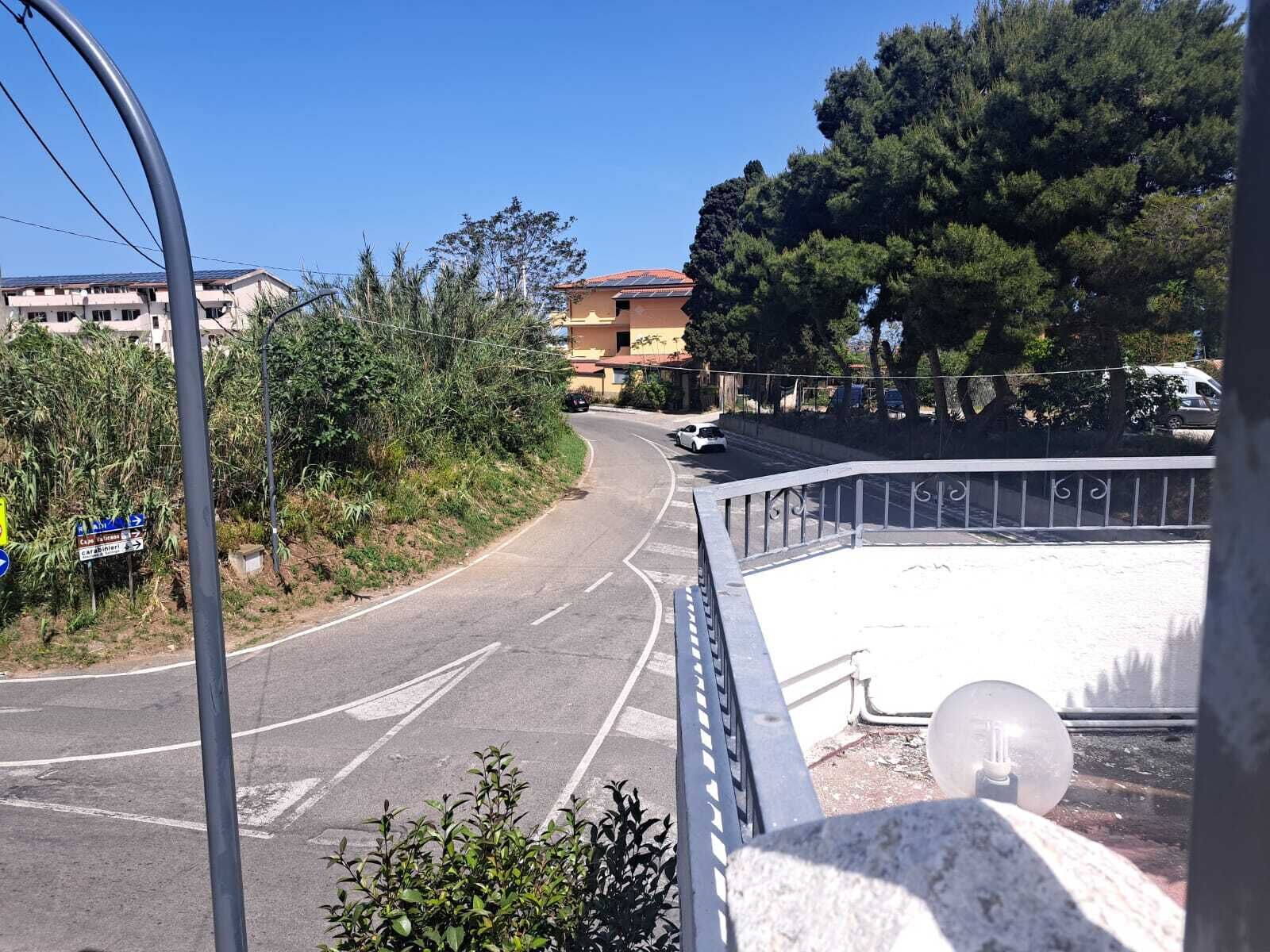 A road curving to the right with a building in the background, surrounded by trees and vegetation.