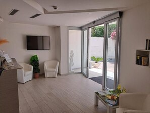 Una sala de estar moderna con un televisor de pantalla plana, dos sillones blancos, una mesa de centro y un amplio ventanal corredero que da a un espacio exterior con plantas.