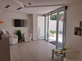 A modern living room with a flat-screen TV, two white armchairs, a coffee table, and a large sliding glass door leading to an outdoor area with plants.