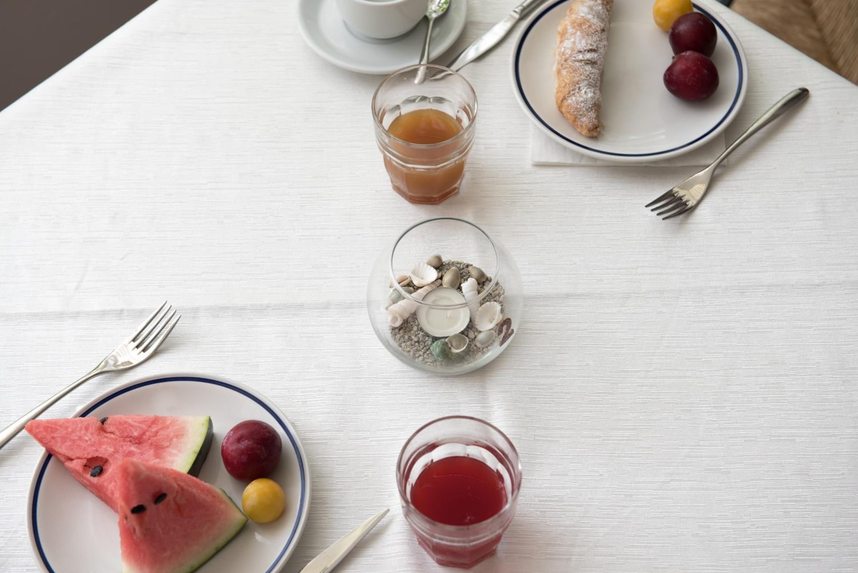 Une table avec une assiette de pastèque, des raisins et un verre de boisson rouge ; une autre assiette avec une pâtisserie et des fruits ; un verre de jus d’orange ; et un petit plat contenant des noix et des graines.