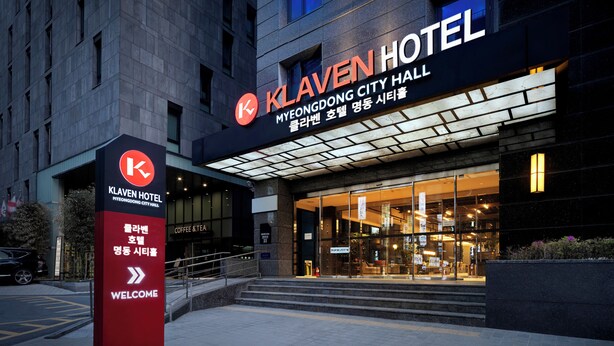 The exterior of Klaven Hotel, featuring a prominent entrance with glass doors, a red sign with the hotel's name, and a welcoming sign.