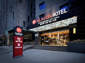 Exterior del Hotel Klaven, destacando una entrada principal con puertas de vidrio, un letrero rojo con el nombre del hotel y una señal de bienvenida.