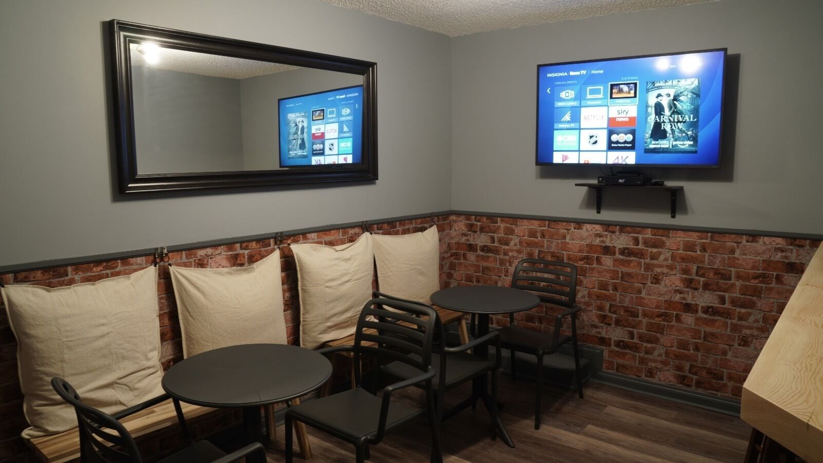 A room with a brick wall, a TV mounted on the wall, a mirror, and a seating area with a table and chairs.