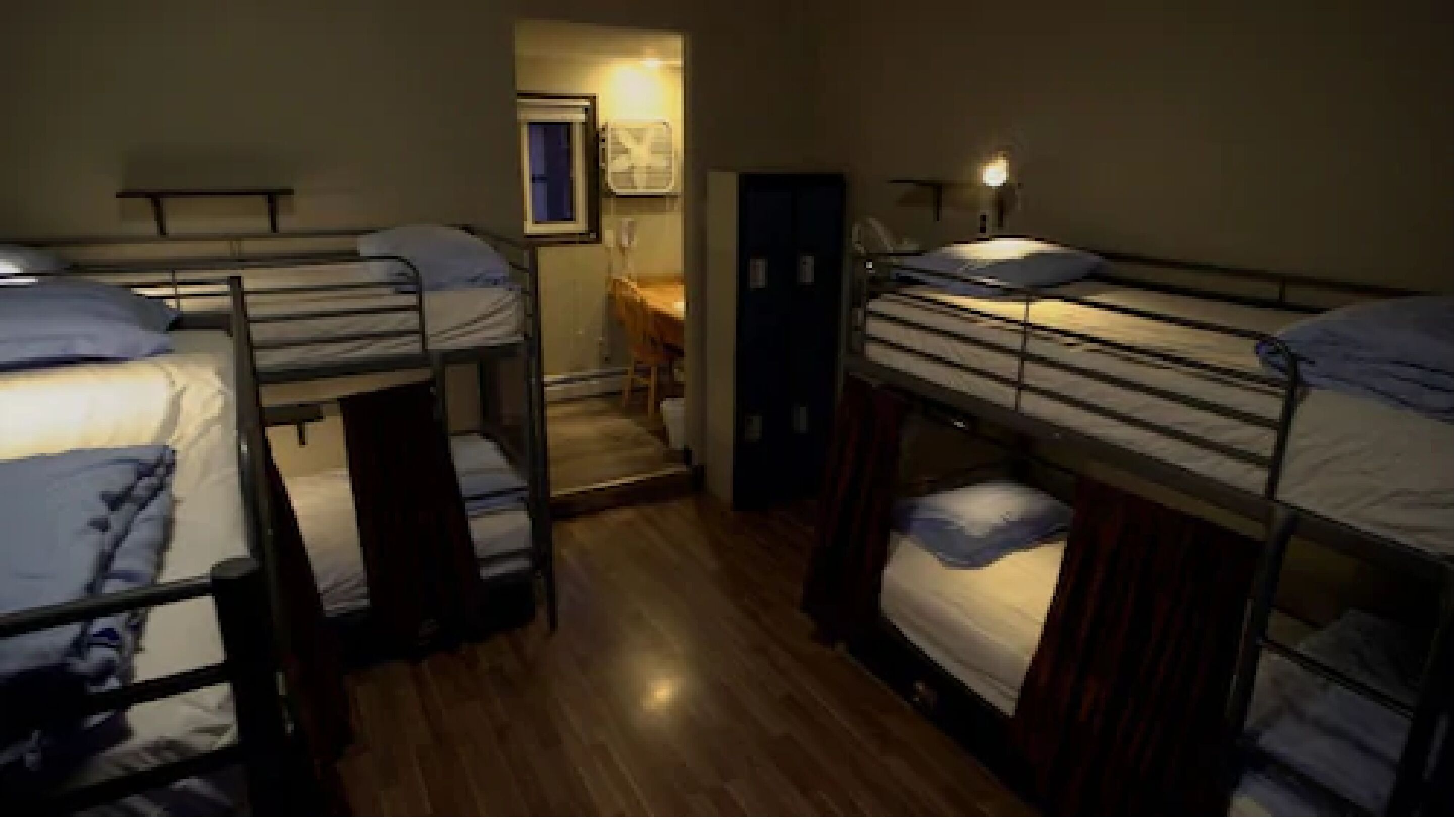 A dormitory room with bunk beds, wooden flooring, a small desk, and a door leading to a bathroom.