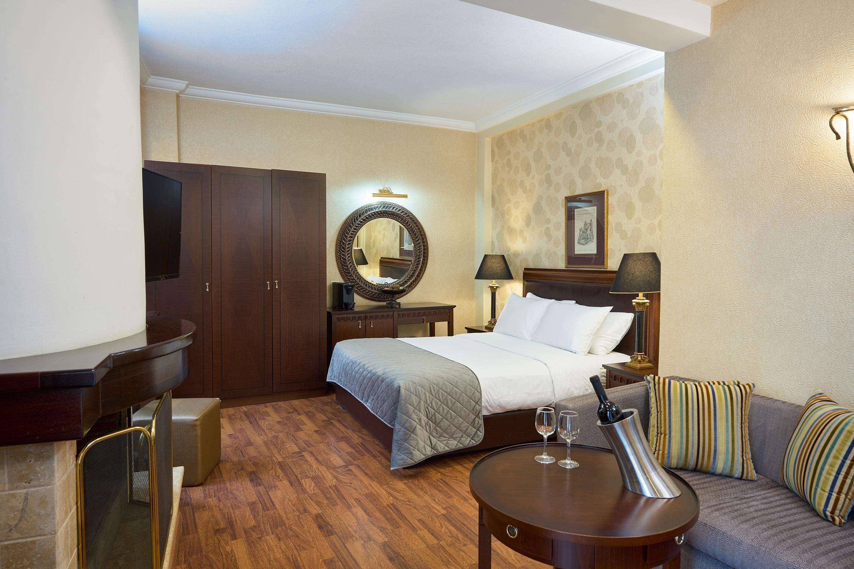A hotel room with a bed, a sofa, a round table, a TV, a mirror, and a fireplace.