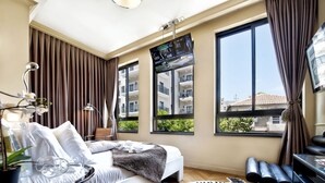 Een moderne hotelkamer met een groot bed, een aan de muur bevestigde televisie, een bureau met stoel, en grote ramen met uitzicht op de stad.