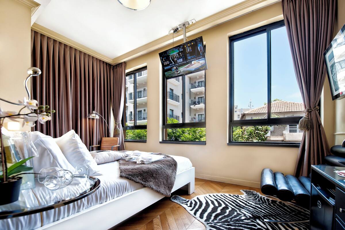 Een moderne hotelkamer met een groot bed, een aan de muur bevestigde televisie, een bureau met stoel, en grote ramen met uitzicht op de stad.