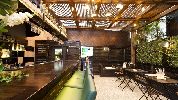 Een barruimte met een houten plafond, groene gestoffeerde zitplaatsen en een aan de muur bevestigd televisiescherm.