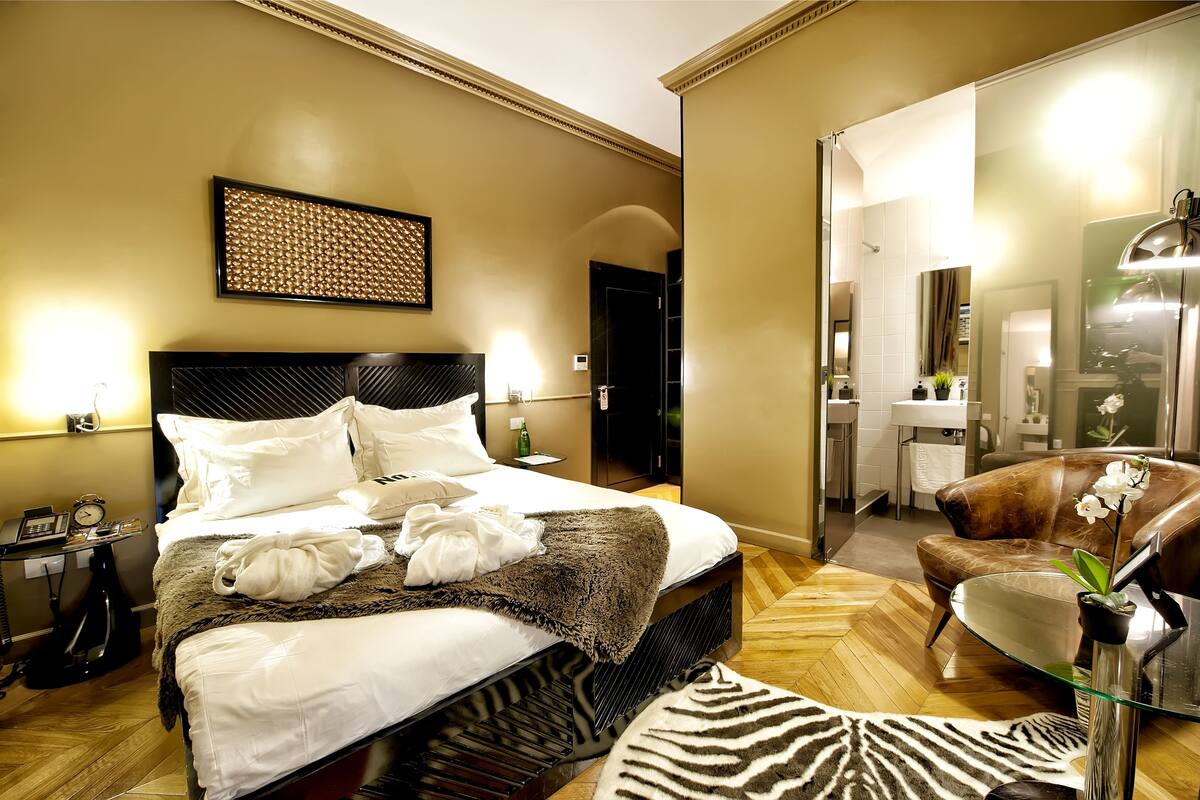 Een hotelkamer met een groot bed, een zithoek met een lederen fauteuil, een glazen salontafel, een vloerkleed met zebraprint en een spiegel die de badkamer weerspiegelt.