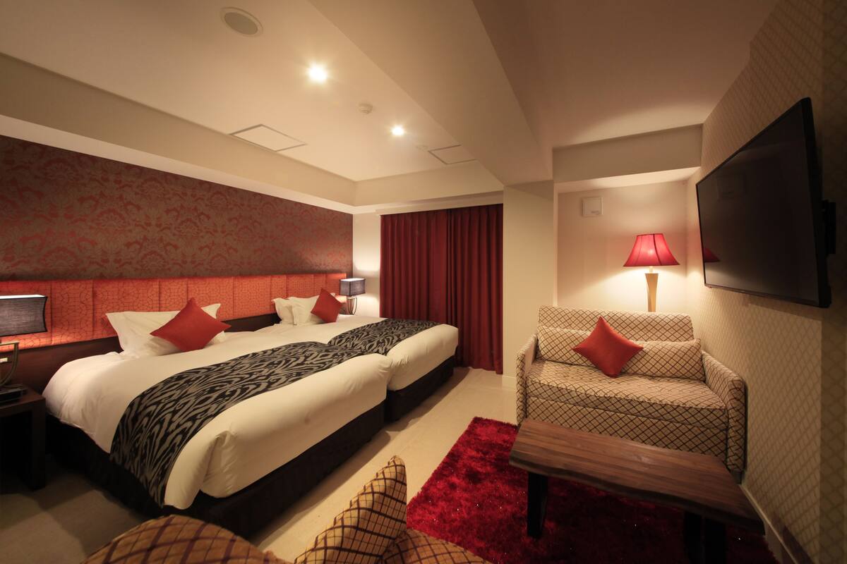 A hotel room with a large bed, two bedside tables, a TV, a red carpet, and a patterned armchair.