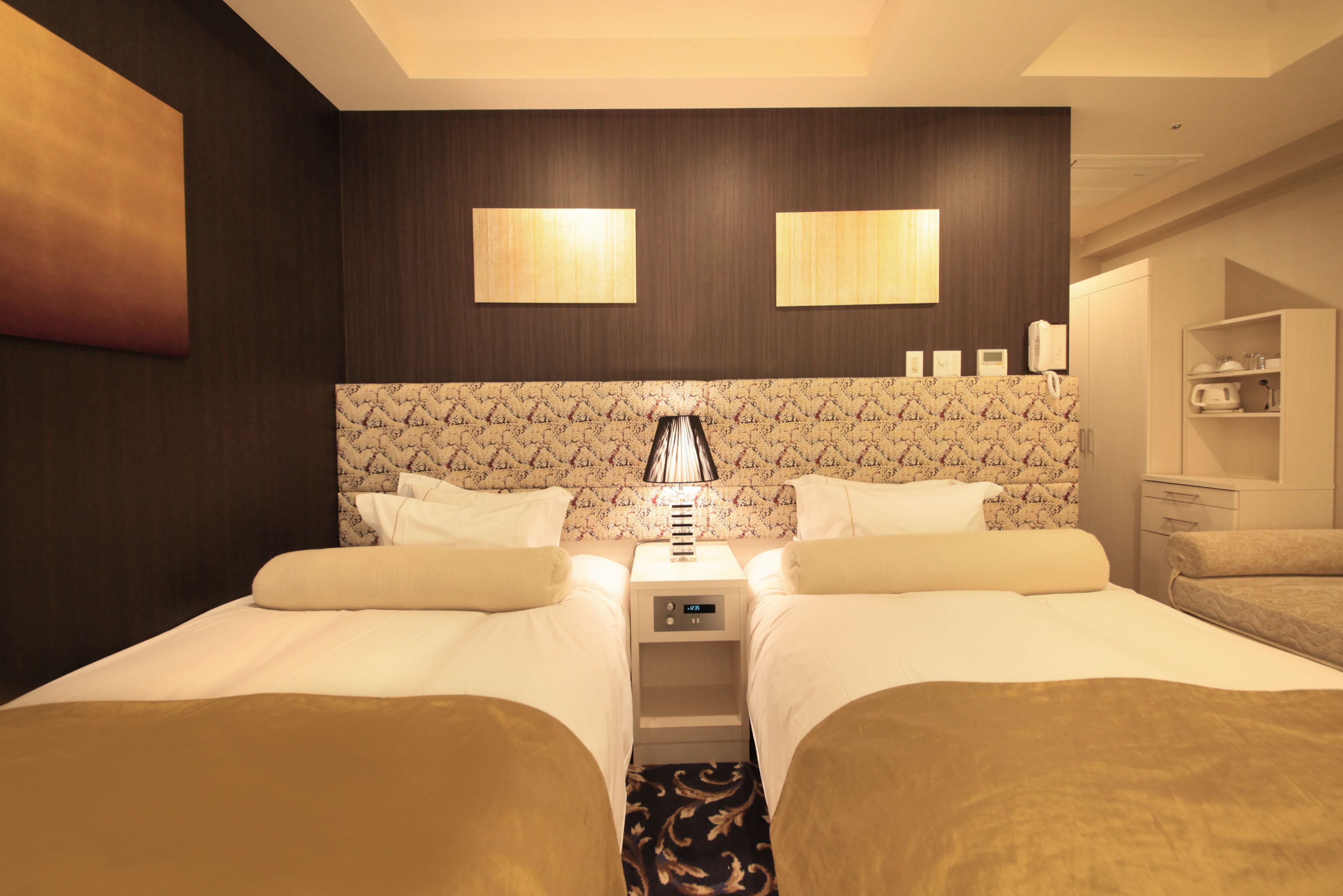 A hotel room with two beds, a nightstand with a lamp, a small shelf with toiletries, and a wardrobe.