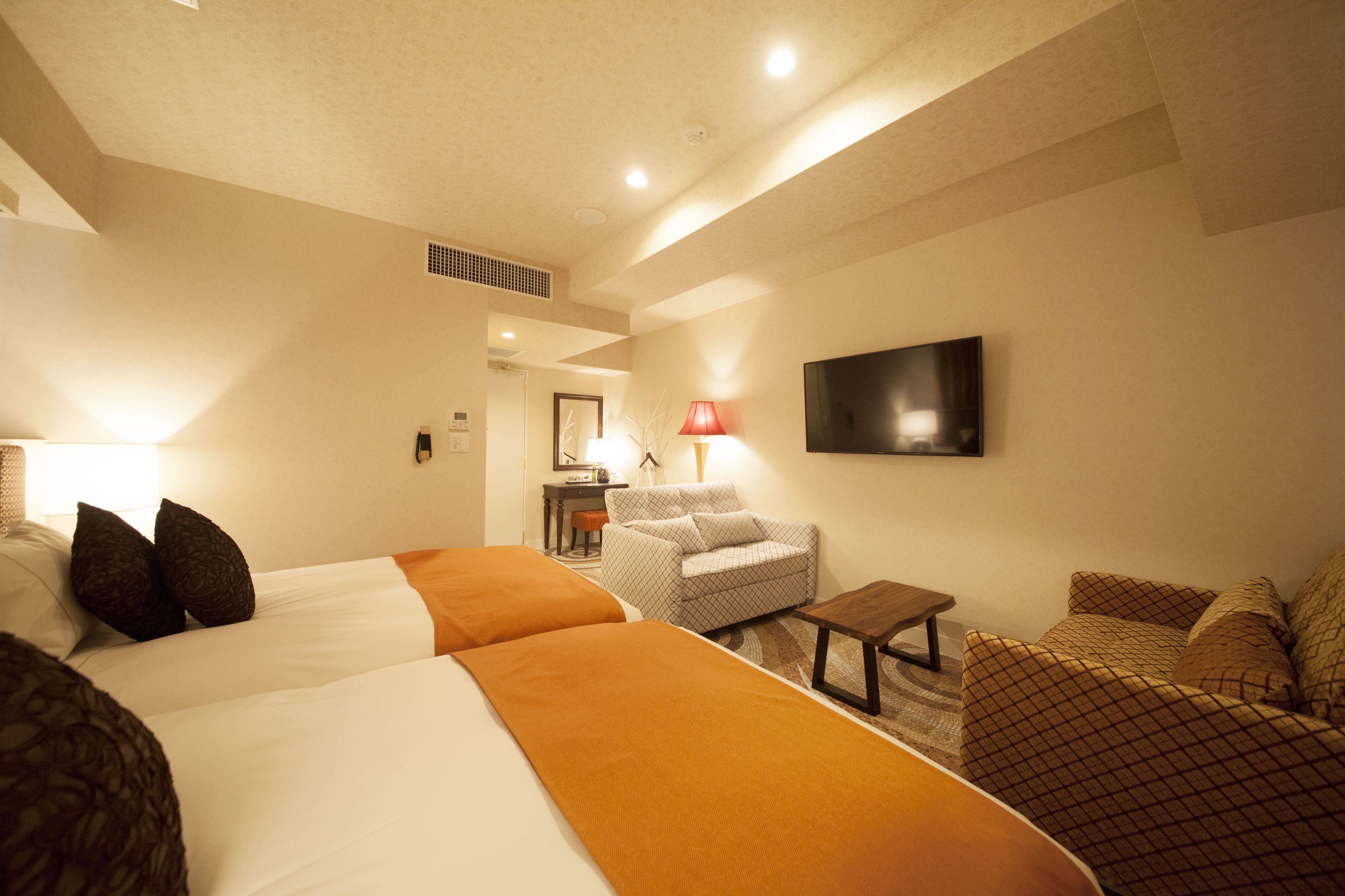 A hotel room with two beds, a sofa, a small table, a lamp, and a TV on the wall.