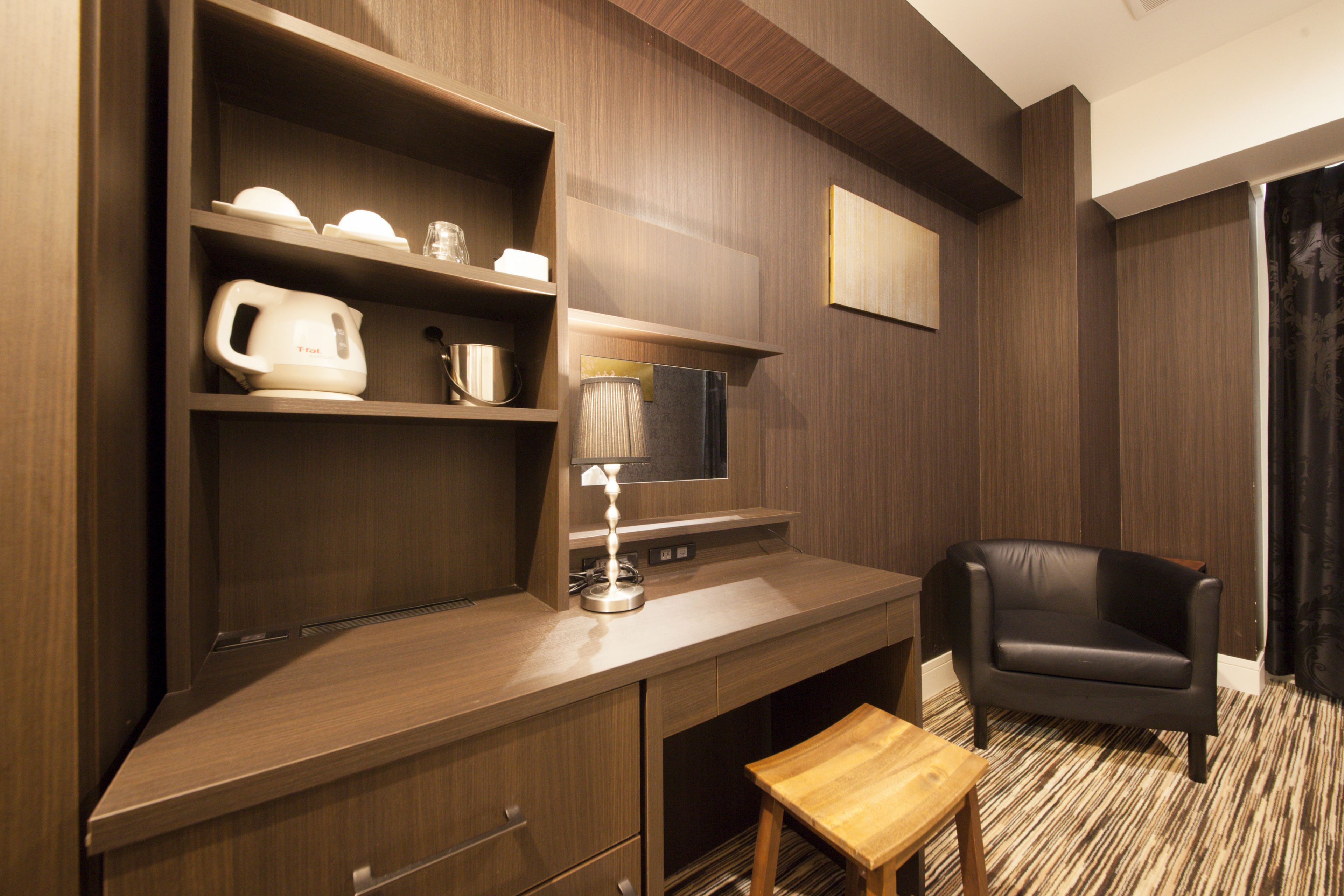 Una habitación de hotel moderna con un escritorio de madera, una silla de cuero negro, un taburete pequeño de madera, un televisor de pantalla plana y una estantería con una tetera y tazas.
