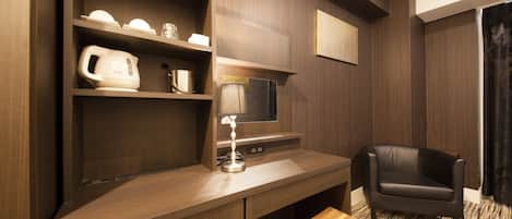 A modern hotel room with a wooden desk, a black leather chair, a small wooden stool, a flat-screen TV, and a shelf with a kettle and cups.