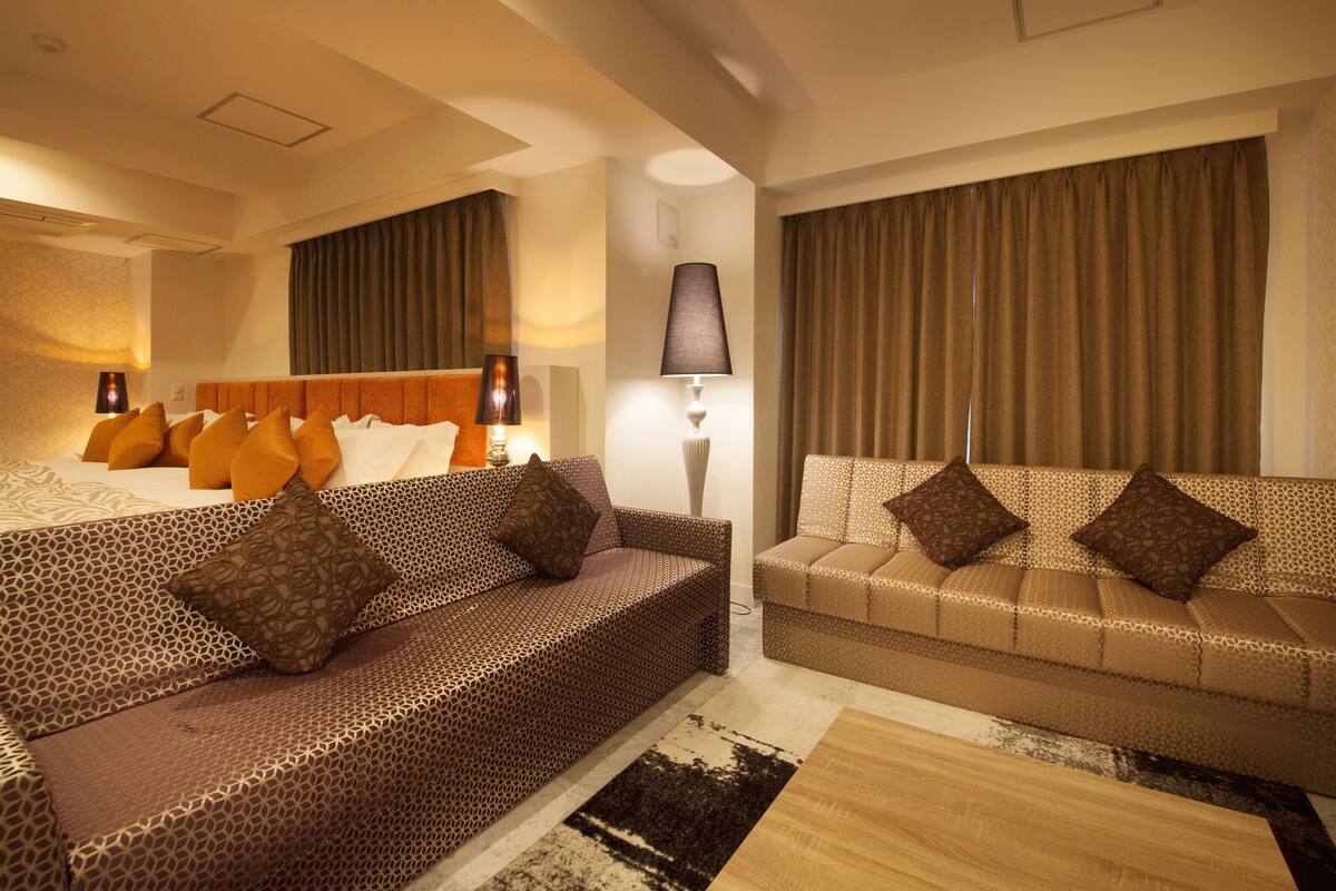 A hotel room with a patterned sofa, a matching armchair, a large bed with multiple pillows, and a floor lamp.