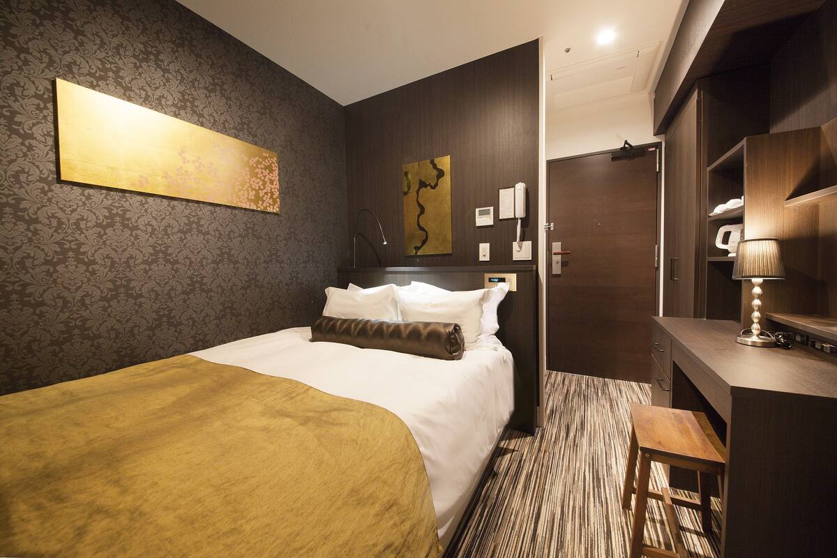 A hotel room with a large bed, a desk with a lamp, a telephone, and a wall-mounted artwork.