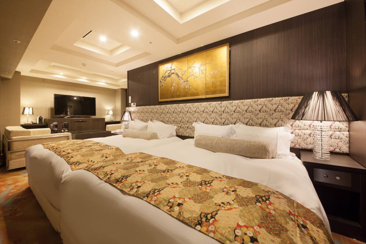 A spacious hotel room with a large bed, a flat-screen TV, a desk with a lamp, and a decorative wall piece.