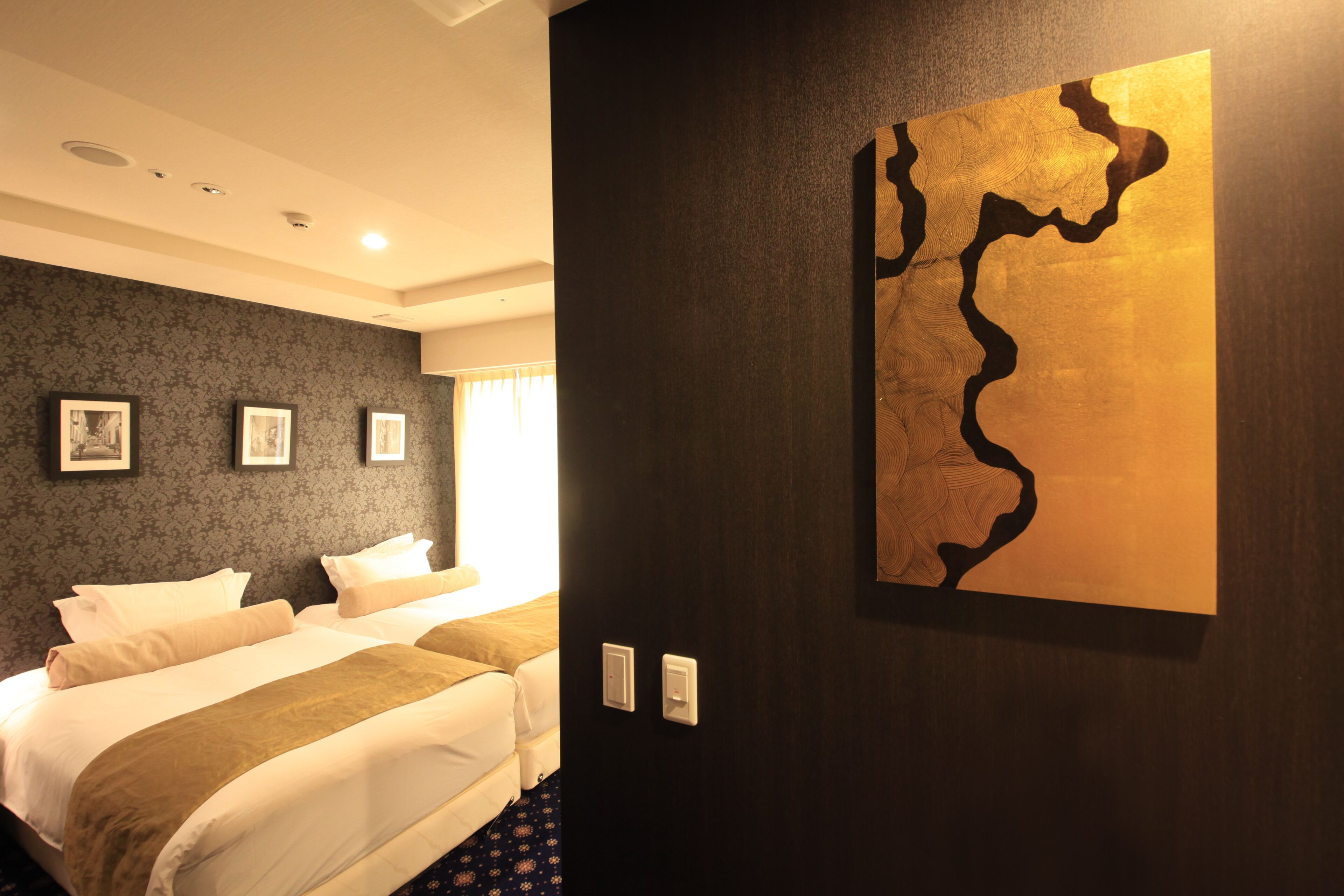 A hotel room with a large bed, a dark wall with a gold abstract painting, and a patterned wallpaper.